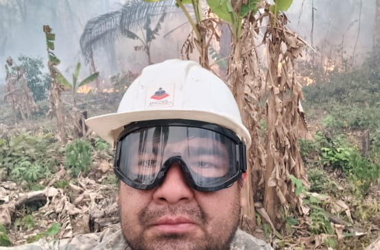 Homem de capacete e óculos de proteção olhando para a câmara, enquanto ao fundo, há arbustos queimando e fumaça subindo