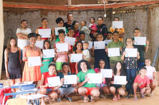 31 pessoas, entre mulheres, homens, jovens e crianças estão em pé, algumas agachadas, em uma sala com paredes brancas ao fundo. A maioria das pessoas segura diante do peito, com as duas mãos, um diploma de conclusão da oficina escrito em folha branca.