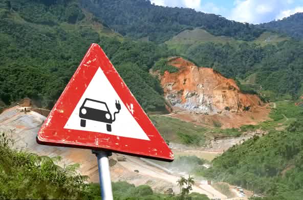 (Montagem de foto) Placa de trânsito com carro elétrico em frente de uma mina de lavra a céu aberto