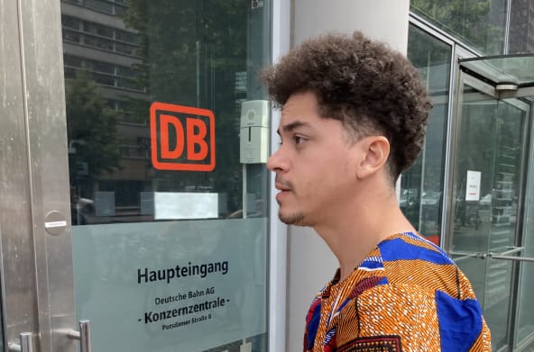 Um jovem está parado com um envelope em frente a uma porta de vidro com a etiqueta DB Entrada principal da sede do Grupo