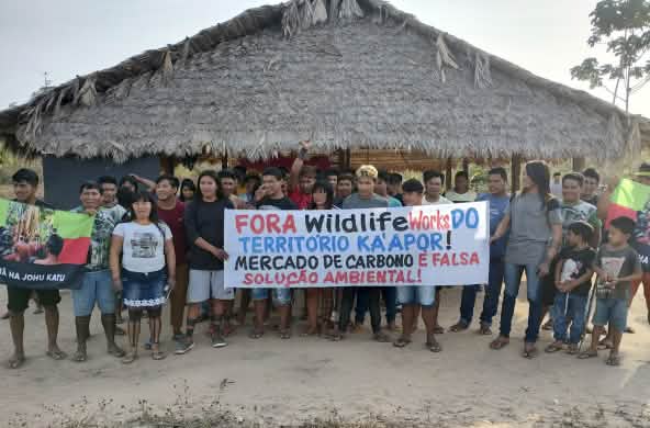 Mais de 30 pessoas do povo Ka'apor estão em pé, diante de uma casa com pilares de madeira e telhado de palha. As pessoas seguram uma faixa de protesto com a frase: "Fora Wildlife Works do território Ka'apor! Mercado de carbono é falsa solução ambiental!"