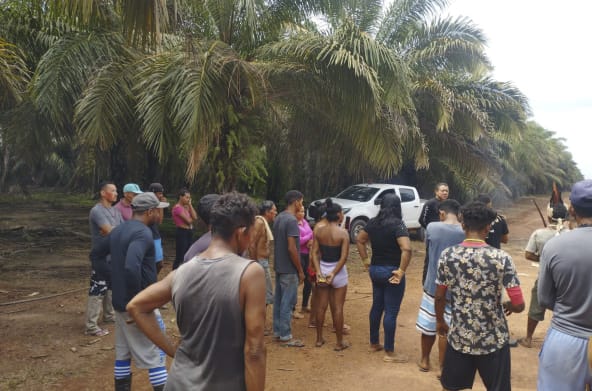 Mais de 15 pessoas indígenas, entre mulheres e homens, aparecem de costas, em pé, em uma estrada de terra. Estão conversando com seguranças privados da empresa Agropalma, que impedem sua passagem