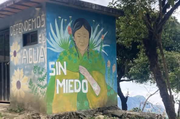 Pintura colorida com plantas e uma mulher indígena sobre o muro de uma pequena edificação, com a inscrição: ""Defendemos a água sem medo"