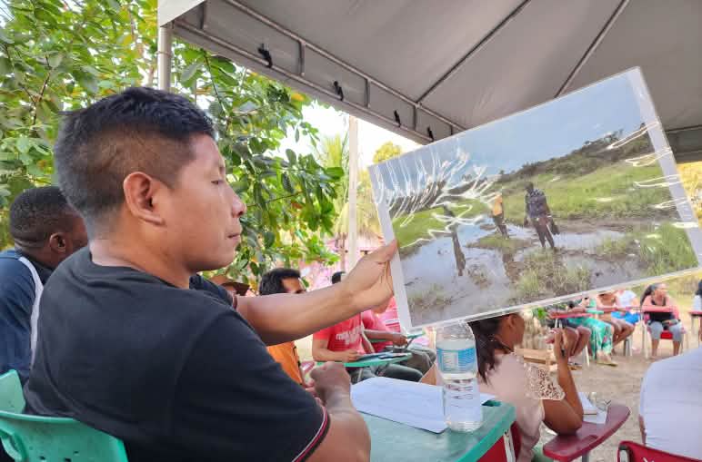Professores Awa Guajá durante o Encontro de Atingidas e Atingidos pela Mineração de 2024, durante conversa com representantes de comunidades da Guiné, África, impactados pela Mina de Simandou Homem Awa Guajá sentado observa uma foto. Ele está em uma cadeira em meio a um círculo com outras pessoas também sentadas