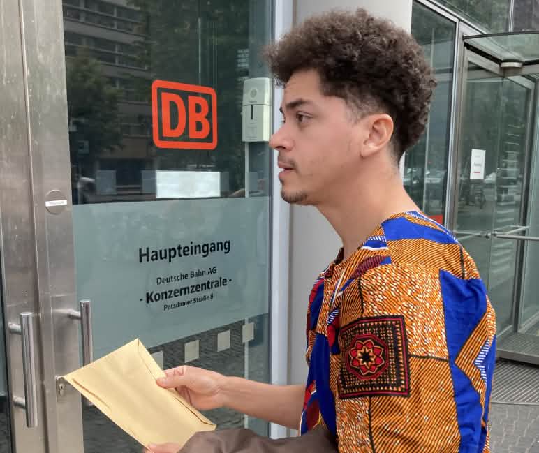 Entrega da carta de reclamação à sede do Grupo DB pelo Justica nos Trilhos Um jovem está parado com um envelope em frente a uma porta de vidro com a etiqueta DB Entrada principal da sede do Grupo