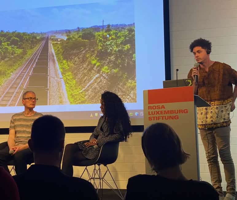 Evento de discussão com Flavia Nascimento e Mikaell Carvalho na Fundação Rosa Luxemburgo em Berlim Três pessoas em um pódio em frente a uma tela com uma foto de vagões de trem em uma linha férrea, na frente deles as cabeças de três espectadores como sombras escuras