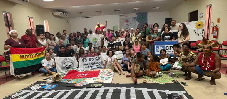 Representantes de povos e comunidades tradicionais participam de seminário sobre potenciais impactos do empreendimento porto-ferroviário Grão-Pará Maranhão, na Amazônia maranhense. Foto de grupo de cerca de 50 pessoas com faixas em uma sala de seminário. Uma das faixas traz o desenho de um trilho de trem.