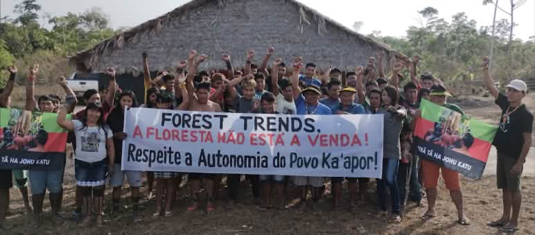 Lideranças do Tuxa Ta Pame se manifestam contra a presença da ONG Forest Trends no território Mais de 30 pessoas do povo Ka'apor estão em pé, diante de uma casa com pilares de madeira e telhado de palha. As pessoas estão com o braço estendido e o punho cerrado. Algumas seguram uma faixa de protesto com a frase: "Forest Trends, a floresta não está