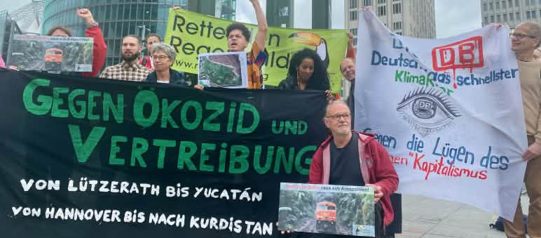 Manifestação na Potsdamer Platz em Berlin, em frente à sede da DB, no fim de maio de 2024 Um grupo de pessoas protesta com grandes faixas coloridas em frente a três torres de escritórios