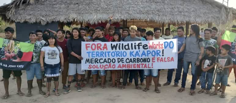 Protesto dos Ka'apor contra negociantes de créditos de carbono em seu território Mensagem escrita na faixa: Fora Wildlife Works do Território Ka'apor! Mercado de créditos de carbono é falsa solução ambiental! Mais de 30 pessoas do povo Ka'apor estão em pé, diante de uma casa com pilares de madeira e telhado de palha. As pessoas seguram uma faixa de protesto com a frase: "Fora Wildlife Works do território Ka'apor! Mercado de carbono é falsa solução ambiental!"