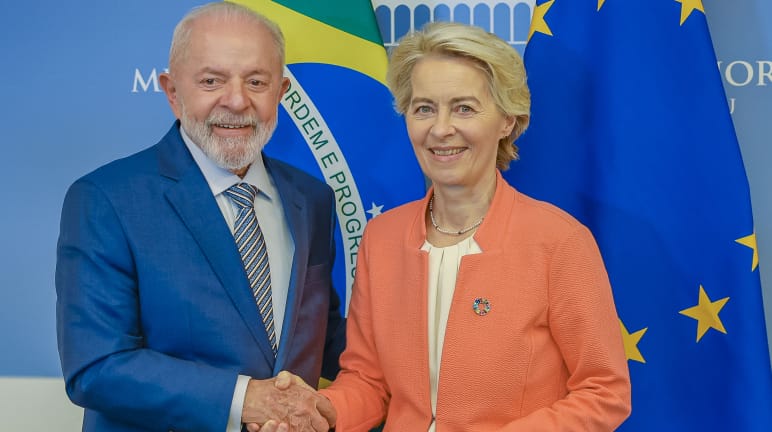 A Presidente da União Europeia, Ursula von der Leyen e o Presidente brasileiro, Lula, apertam-se as mãos diante das bandeiras nacionais