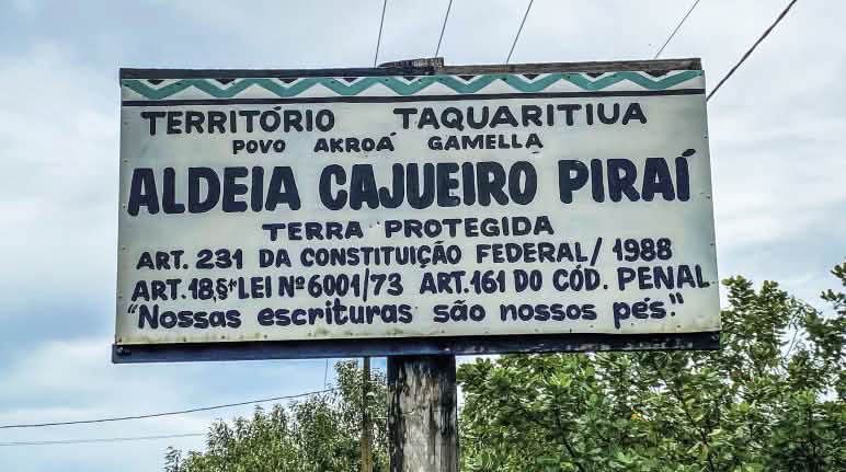 Placa na entrada da Terra Indígena Taquaritiua Placa com fundo branco e letras azuis. Está escrito Aldeia Cajueiro Piraí, terra protegida. Nossas escrituras são nossos pés
