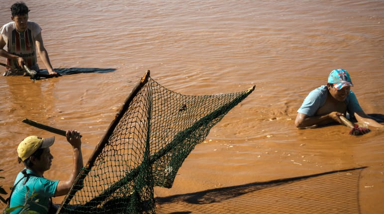 Três homens pescando, em pé, com água até a cintura, em um rio marrom