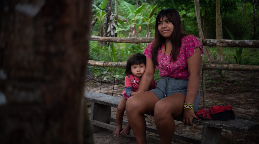 Uma mulher indígena senta-se com uma criança em um banco de madeira na floresta tropical