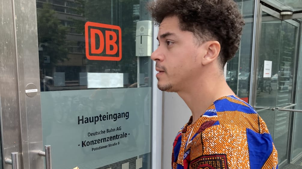 Um jovem está parado com um envelope em frente a uma porta de vidro com a etiqueta DB Entrada principal da sede do Grupo
